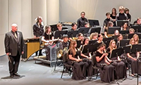  photo of band director dressed in a suit with students holding band instruments sitting nearby